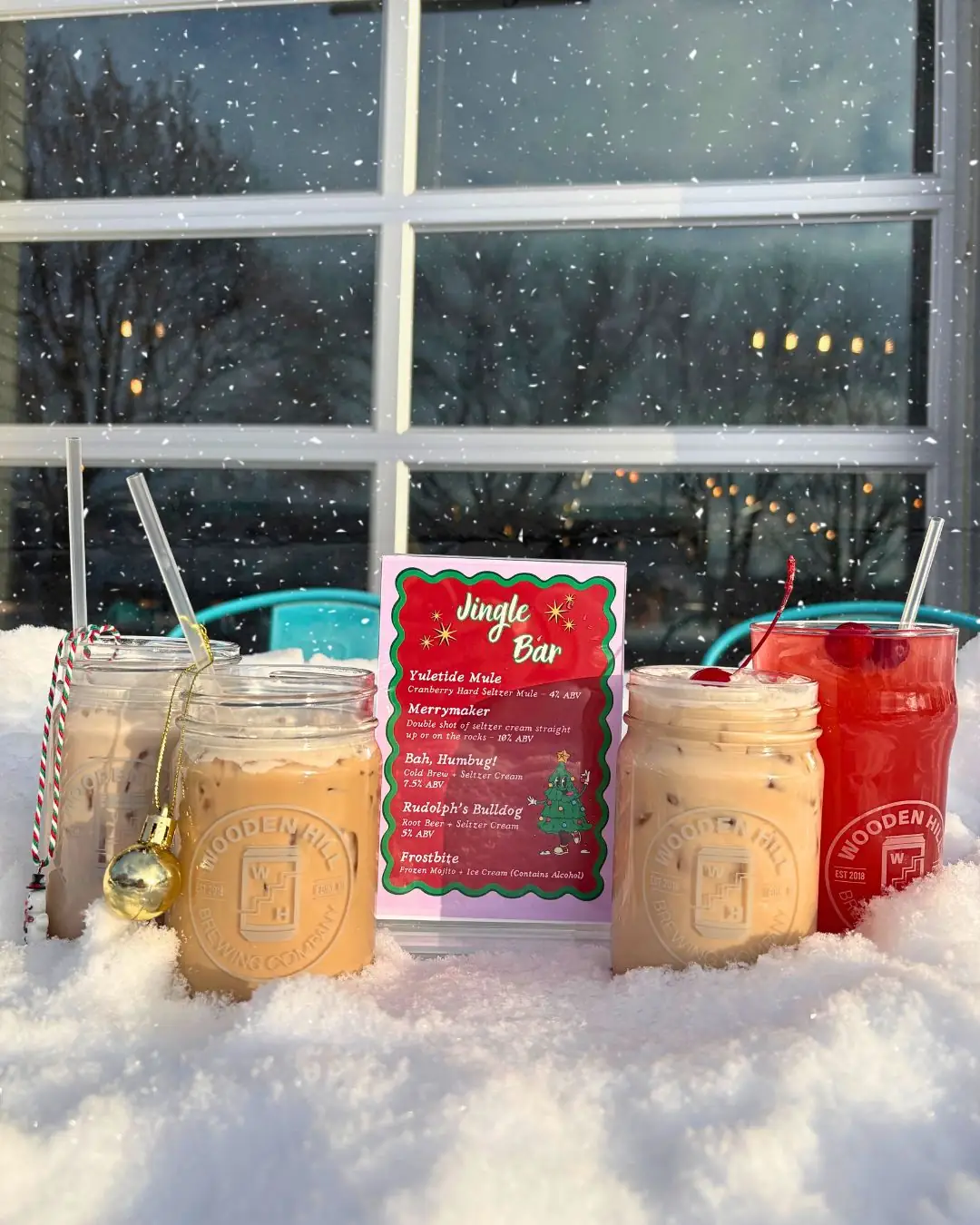 Image of four wooden hill drinks in the snow with jingle bar menu in the center and snow falling in the background. 3 drink are creamy coffee colored in major jars and one is a transparent red.