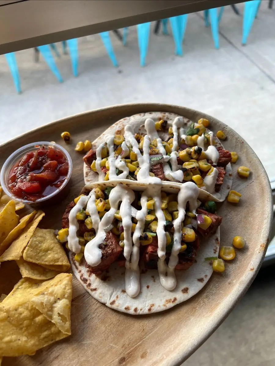 Two steak and elote soft shell tacos with a drizzle of creamy elote sauce and a side of chips and salsa on a wooden plate