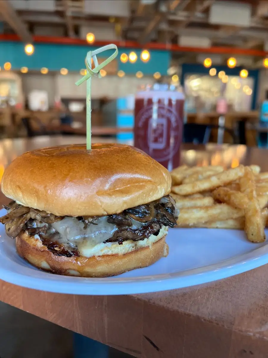 Mushroom and swiss burger on a brioche bun with a side of fries on a white plate. Wooden Hill Beer in background