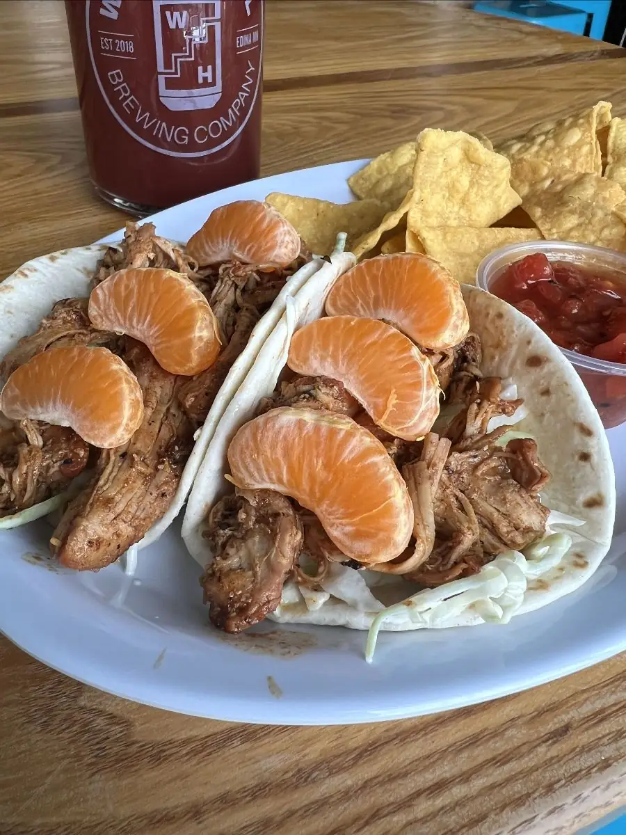 two soft shell tacos with coleslaw, orange marinated chicken, and three mandarin orange slices on top. A side of chips and salsa on the plate and a wooden hill beer in the background.