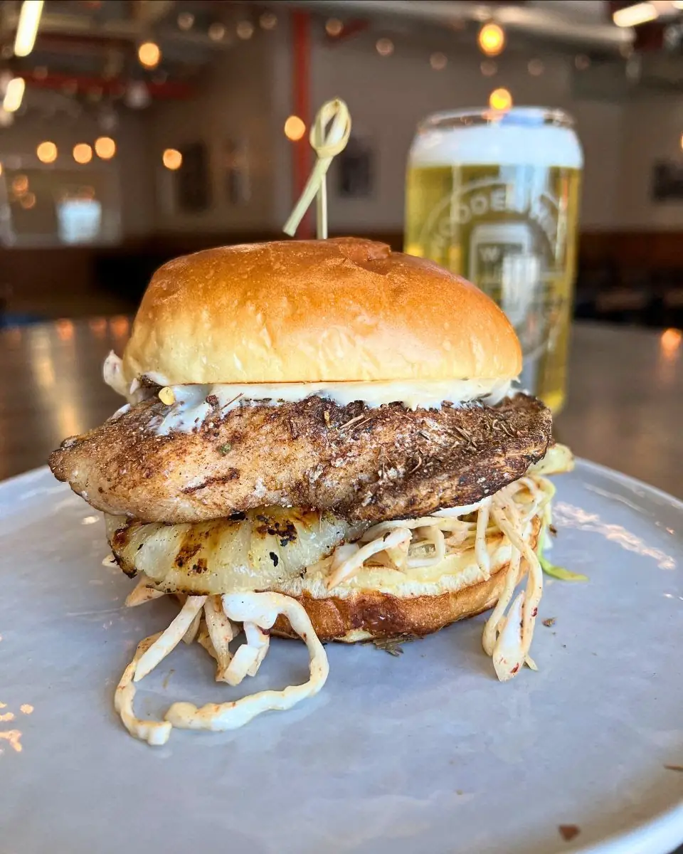 Image of a jerk grilled chicken sandwich on a plate with a Wooden Hill glass of a light beer in the background on the table.