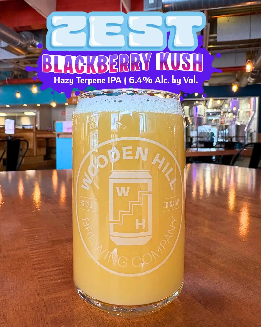 Image of a Wooden Hill glass of a lighter beer with the brewery in the background. Logo at the top that says ZEST Blackberry Kush | Hay Terpene IPA | 6.4% ABV - in blues, purple. pink.