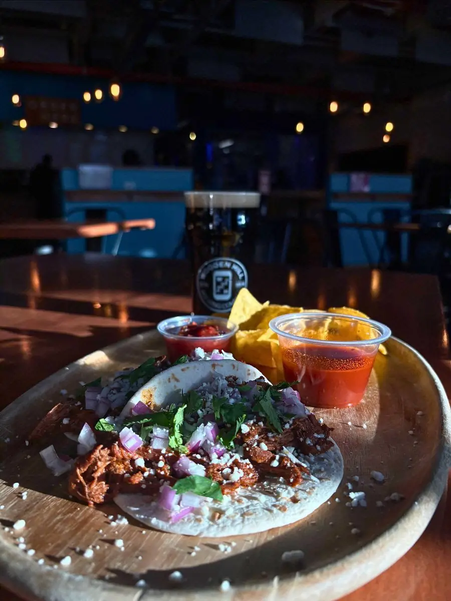 Image of birria tacos on a wood plate with salsa, consomme, and chips on the plate. A glass of a dark beer in the background.
