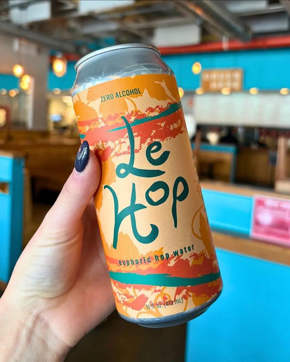 Image of a hand holding a can of Le Hop that has orange and teal designs on it. The background is of the taproom with a lot of blue.