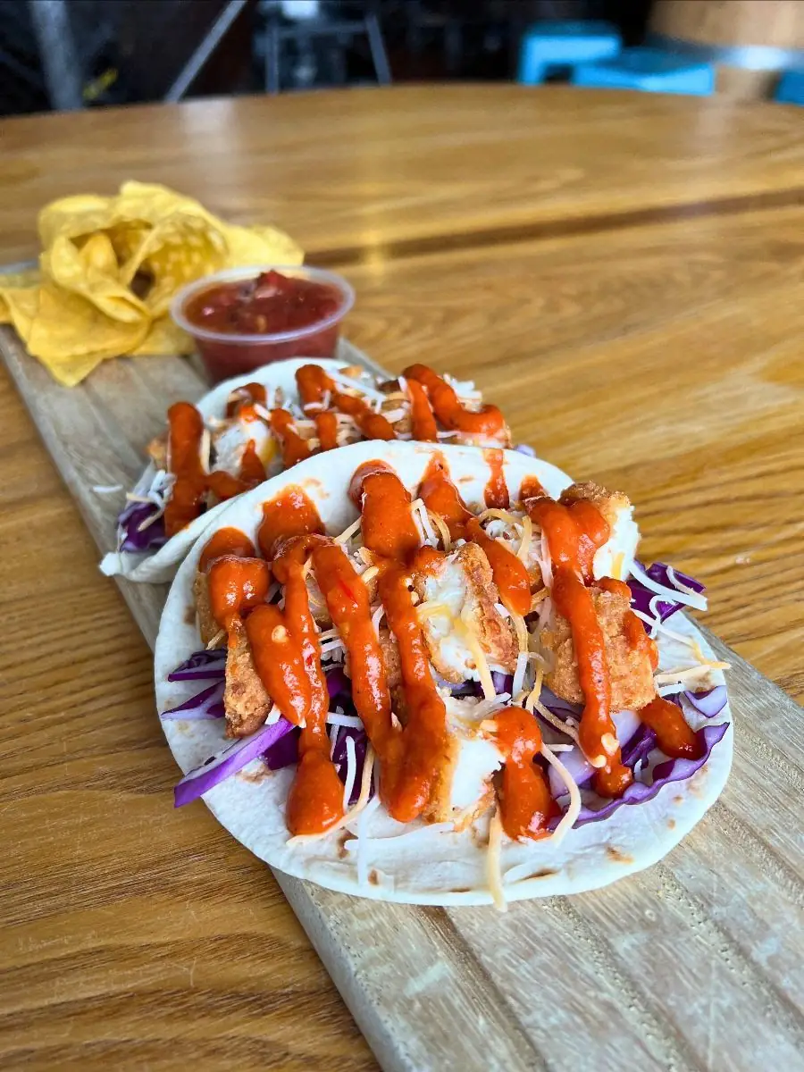 Image of two fiery crispy chicken tacos with chips and salsa on a wooden board on a wood table.