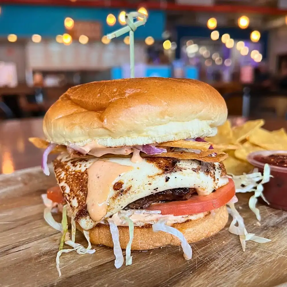 Image of a stacked taco burger on a wood plate with some chips and salsa in the background.