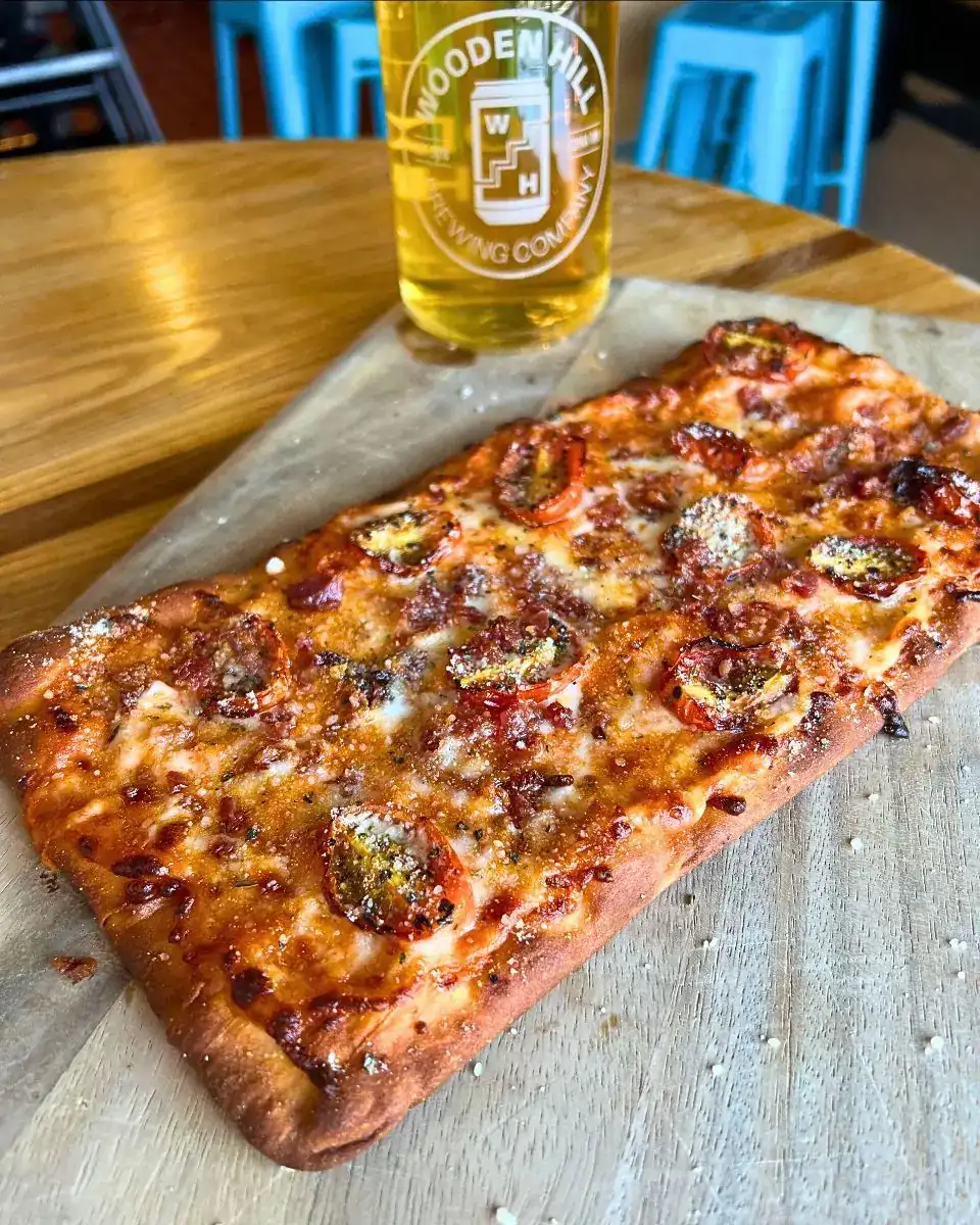 Image of a wood board with spicy vodka bacon flatbread on it and a Wooden hill glass with a light beer in it.