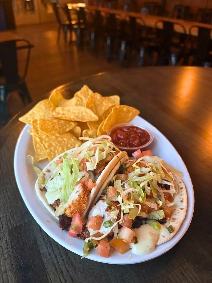 Image of two fiesta potato tacos on a plate with chips and salsa. Tables and chairs in the background.