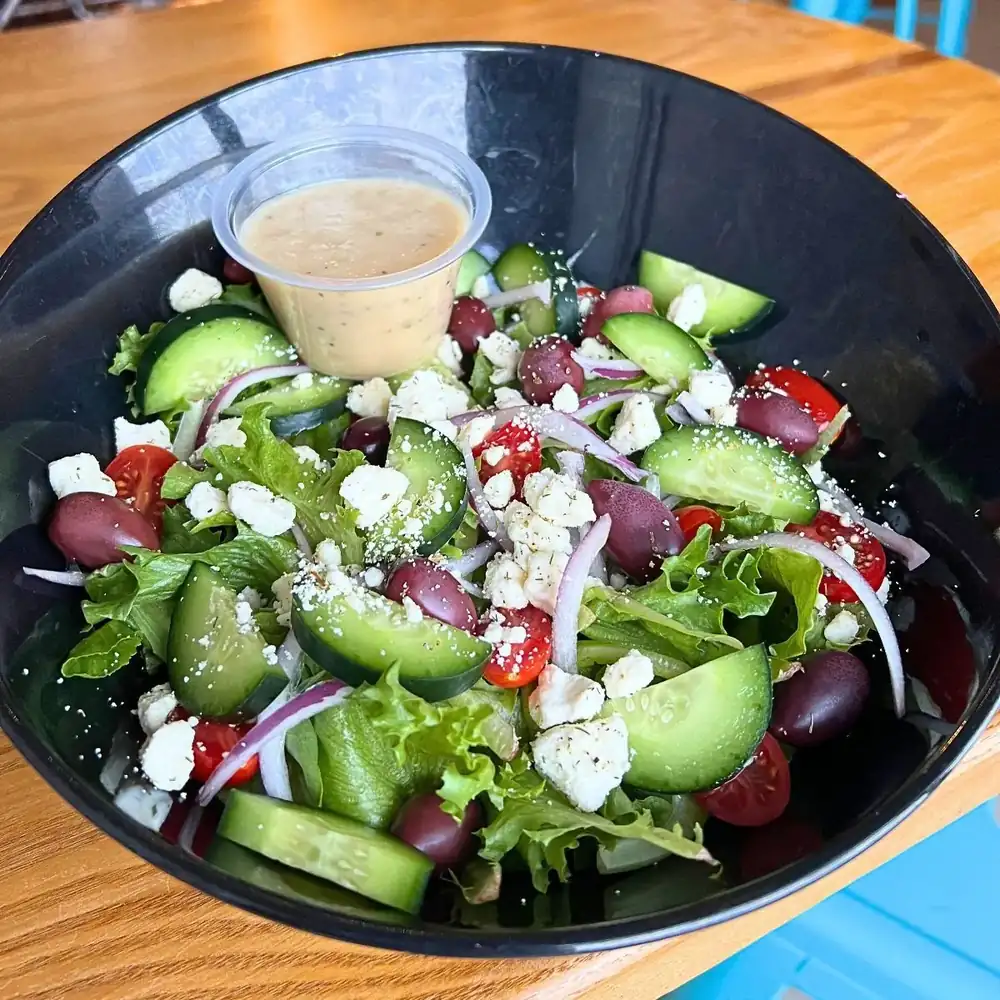 Image of greek salad in a black bowl with a cup of a light colored dressing in it.