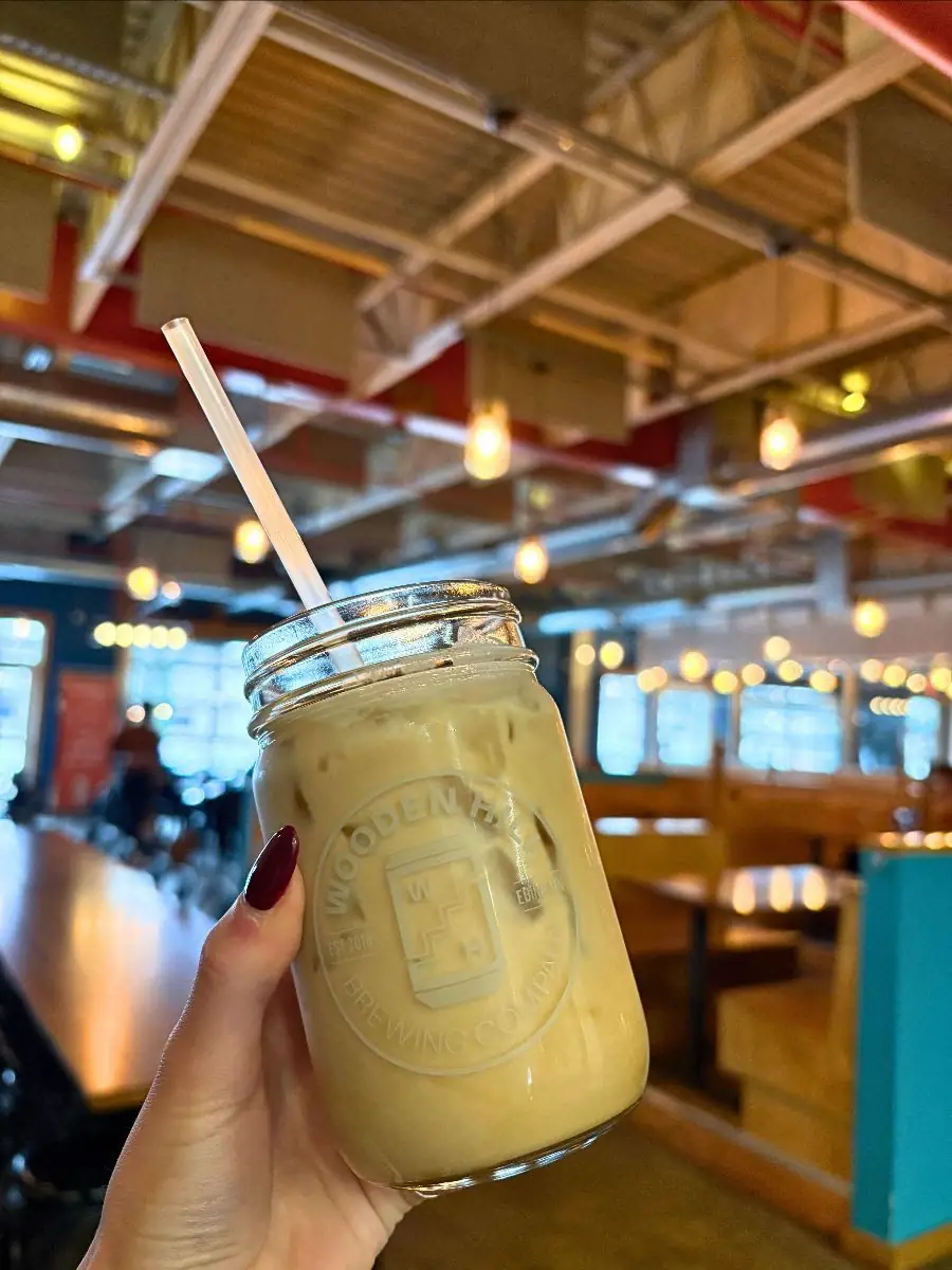 Image of a hand holding a Wooden Hill branded mason jar with a creamy rootbeer beverage in it. Background is of the taproom with tables, chairs, and lights.