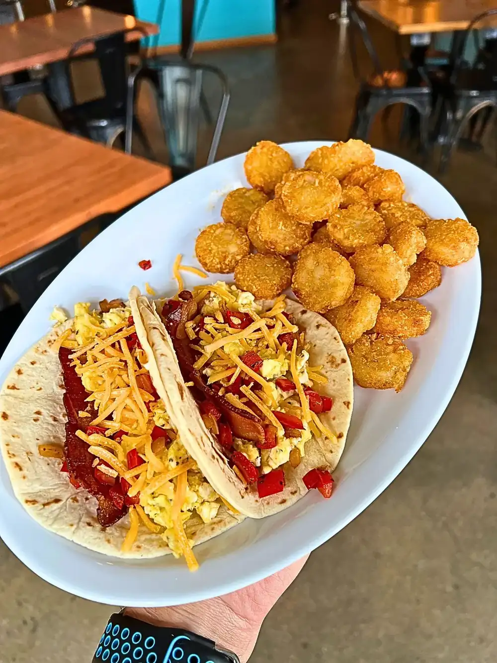 A hand holding a white oval plate featuring two Breakfast Scramble Tacos on soft tortillas. The tacos are loaded with fluffy scrambled eggs, strips of double-smoked bacon, sautéed red peppers and onions, and a generous topping of shredded cheddar cheese. A large side of crispy, golden-brown seasoned tater tots fills the rest of the plate, with blurred taproom tables in the background.