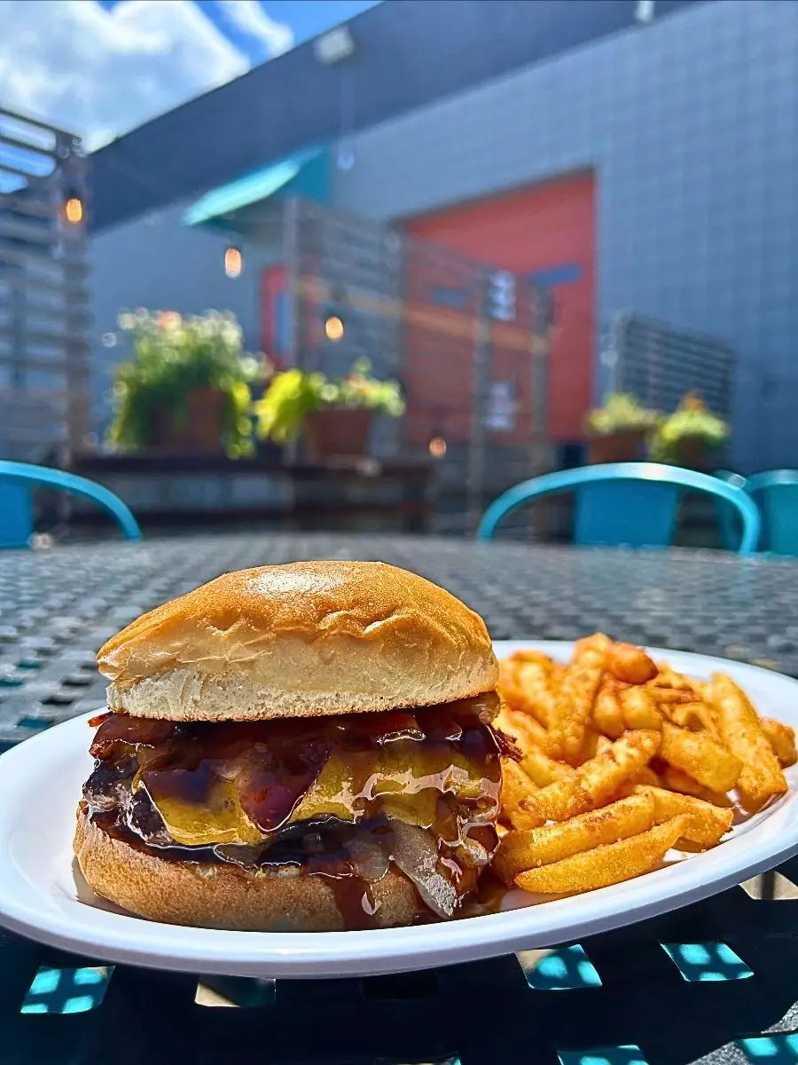 A Bourbon Cheddar Burger featuring a beef patty, yellow cheddar, caramelized onions, bacon, and bourbon sauce on a brioche bun, served with fries on an outdoor patio table.