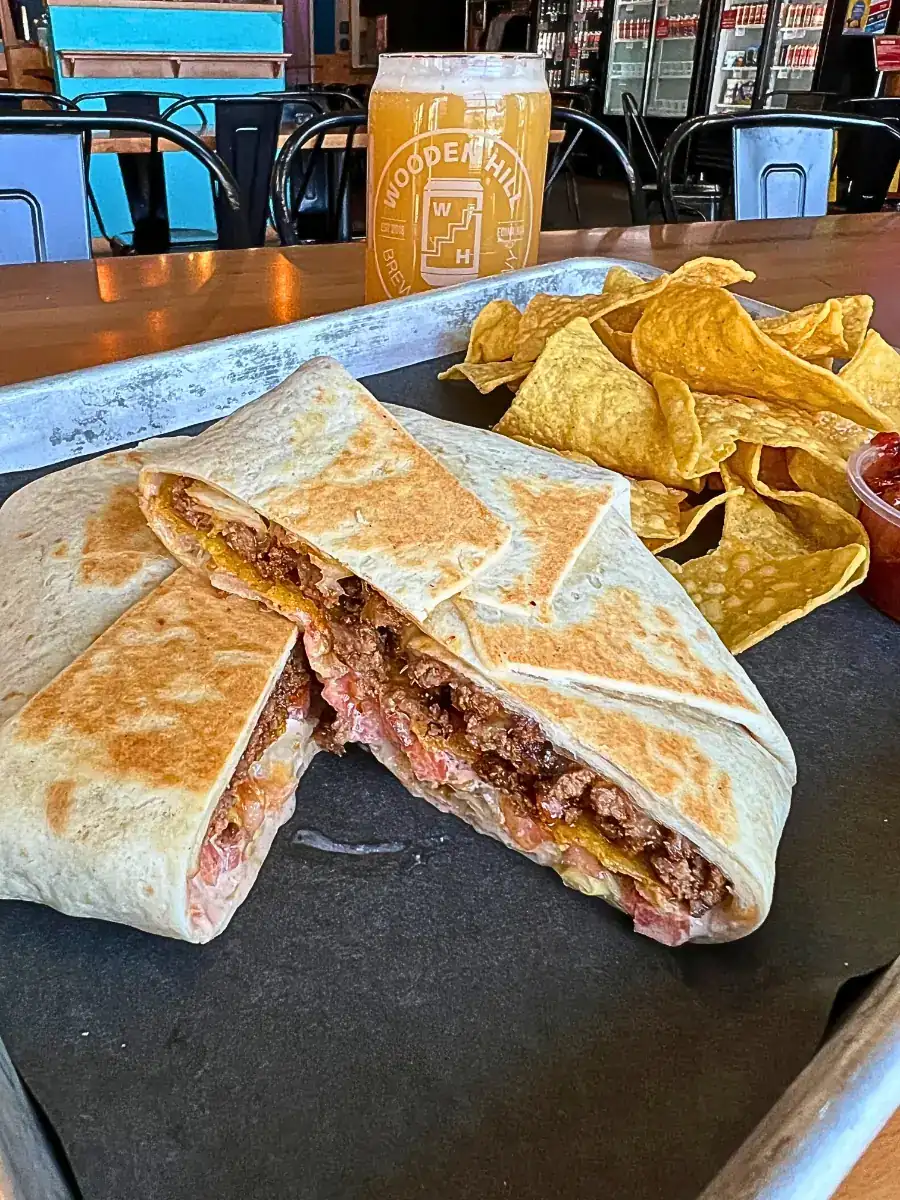 A halved Crunchy Wrap showing delicious layers of seasoned beef, cheese, and fresh veggies, served with a side of chips and a glass of beer.