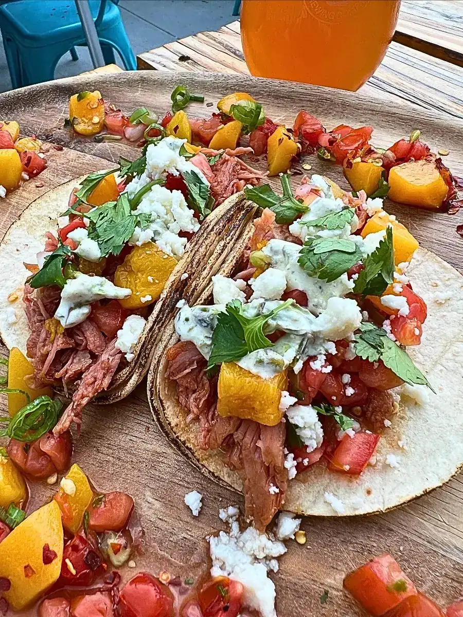 A close-up of two Peach Pork Picante Tacos on a rustic wooden platter. The lightly toasted tortillas are overflowing with slow-cooked shredded pork, bright fresh peach salsa, crumbled white queso fresco, charred jalapeño crema, and fresh cilantro. Extra peach salsa is scattered around the board, with a cold, hazy craft beer visible in the background.