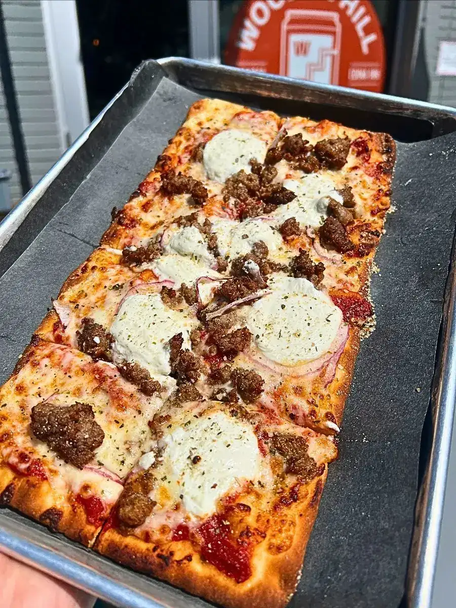 Sausage Ricotta Flatbread: A freshly baked, rectangular flatbread cut into squares and served on a black paper-lined metal tray. The crispy crust is generously topped with house red sauce, melted shredded mozzarella, large dollops of ricotta cheese, crumbled mild sausage, thin slices of red onion, and a sprinkle of house herb and cheese seasoning. A blurred Wooden Hill logo sign is visible in the background.