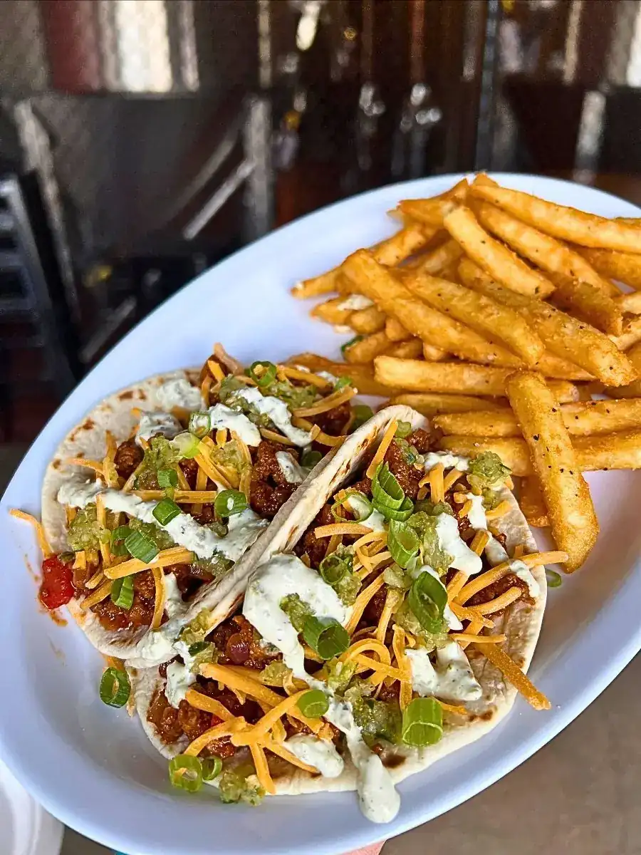 Two Sloppy Joe tacos loaded with beef, cheddar cheese, relish, jalapeño crema, and green onions, served on a white plate with a side of crispy french fries.
