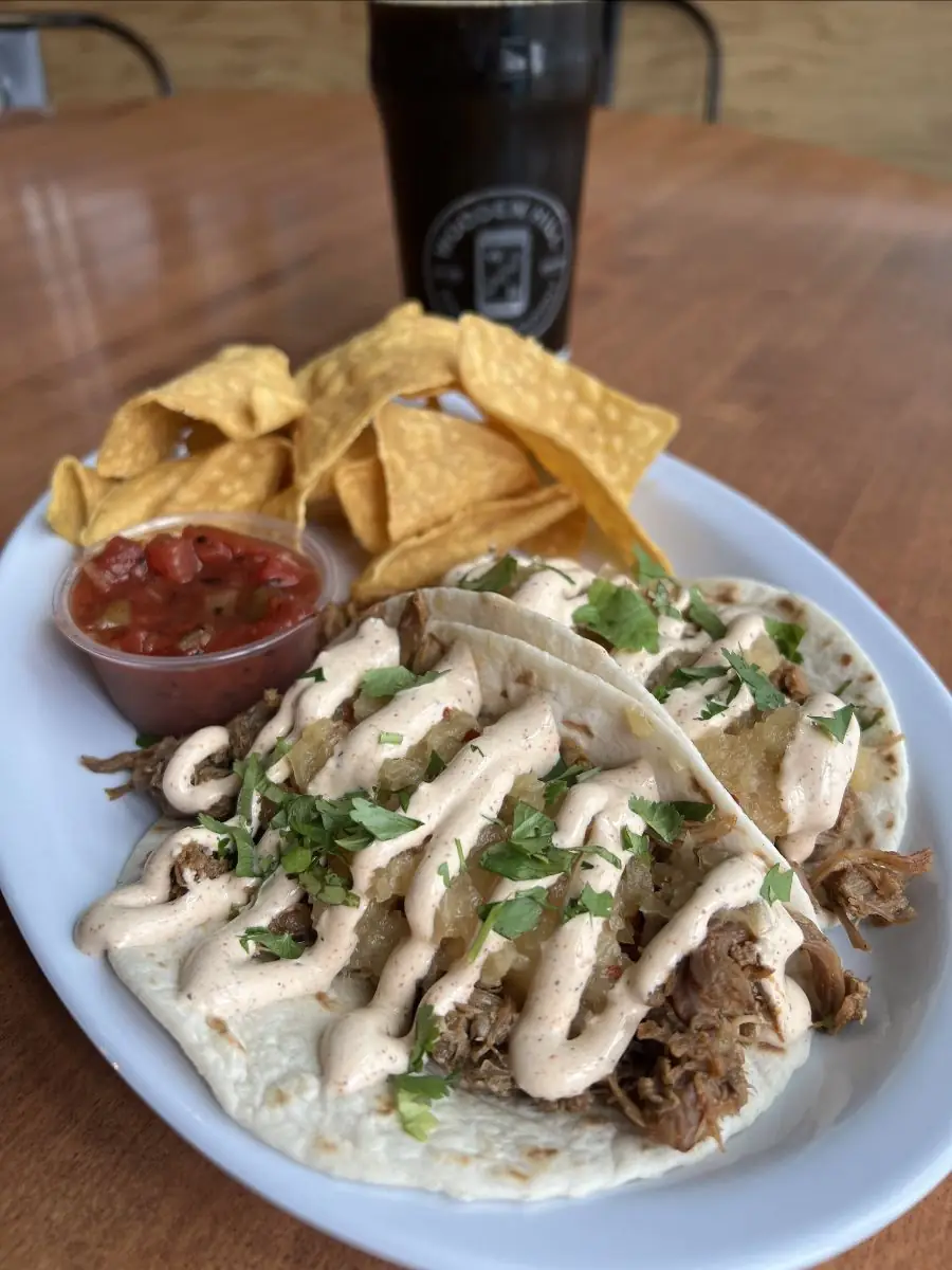 two soft shell tacos with BBQ pulled pork, sweet and spicy pineapple chutney, house seasoned sour cream, and fresh cilantro on top with a side of chips and salsa.