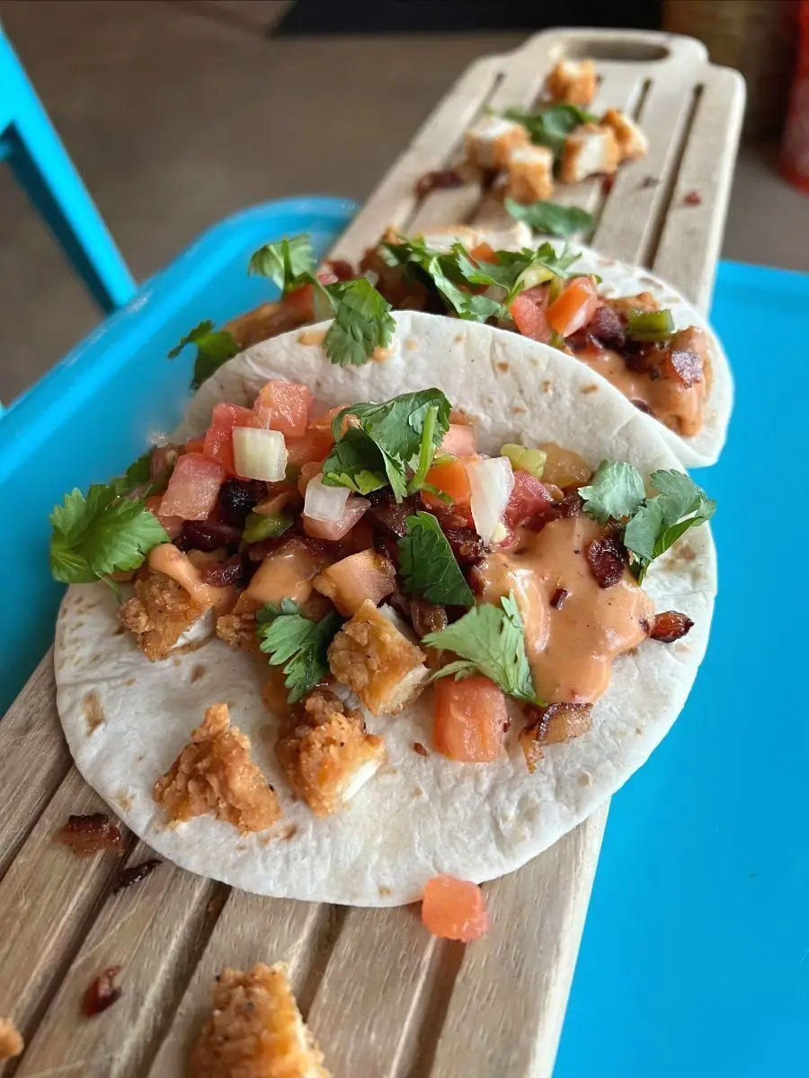 Two crispy chicken tacos on flour tortillas, topped with house creamy queso, crunchy bacon, pico de gallo, and fresh cilantro, resting on a wooden serving board