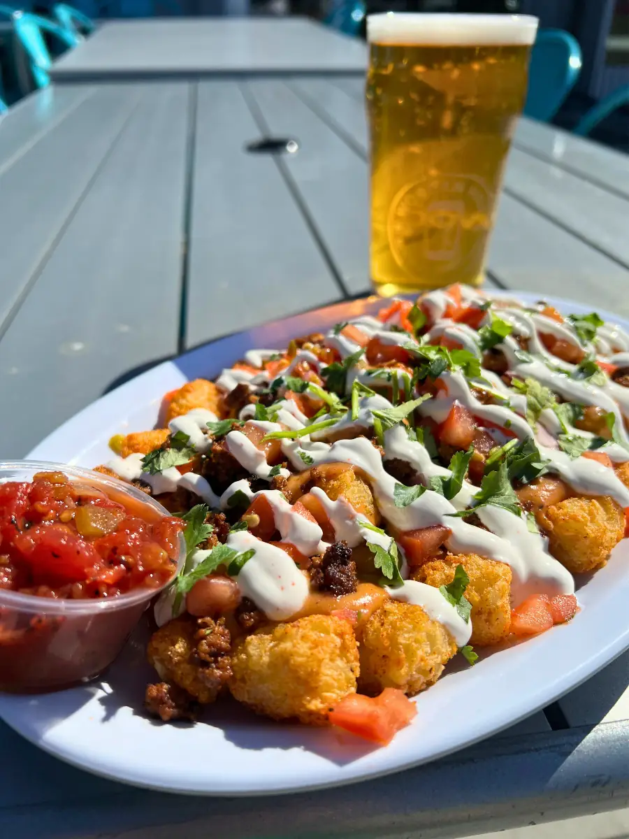 A sunny patio shot of loaded tatchos! Crispy tater tots covered in ground meat, tomatoes, cilantro, sour cream, and queso, served with a side of red salsa. A glass of Wooden Hill beer rests in the background on a grey outdoor table.