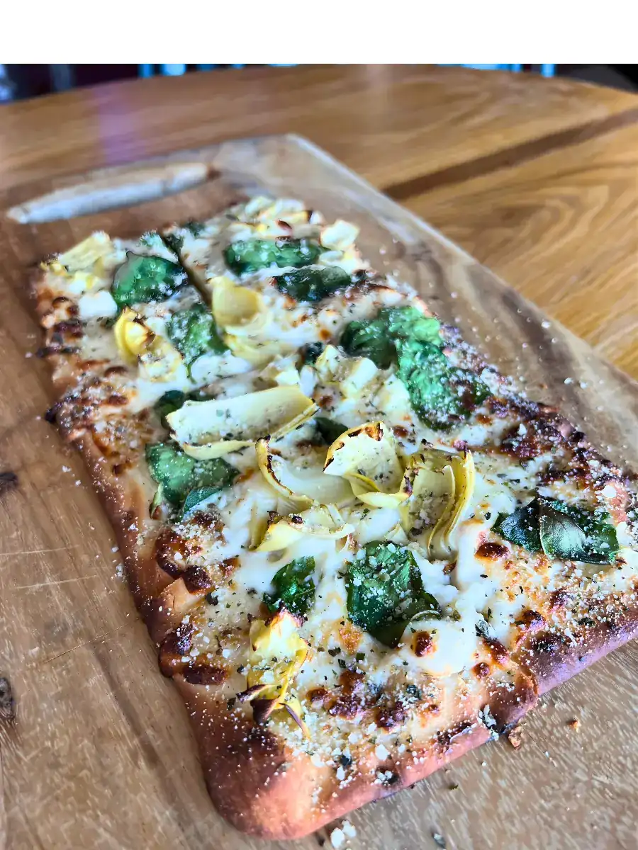 Crispy spinach and artichoke flatbread featuring a creamy white cheese base, fresh spinach, artichoke hearts, and grated parmesan, plated on a wooden cutting board.