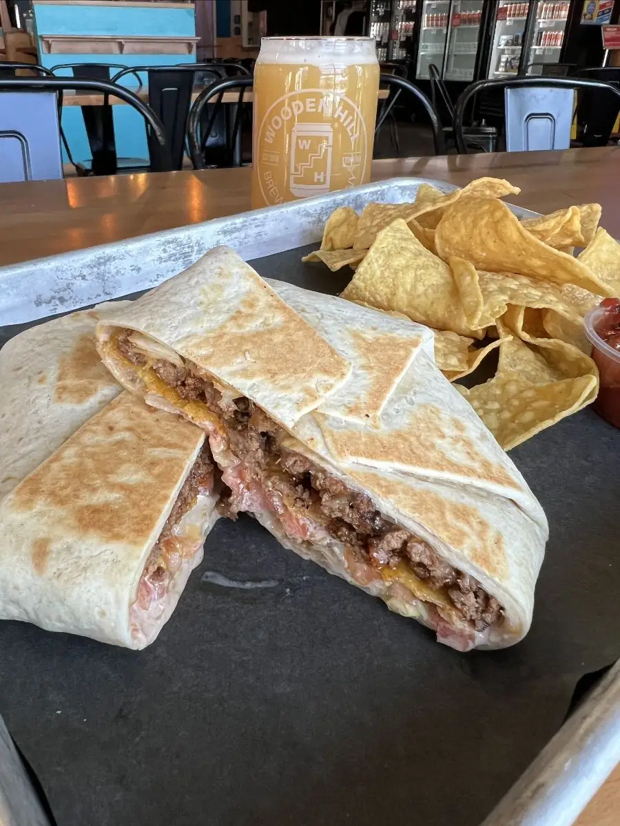 A folded grilled tortilla filled with seasoned ground beef, cheese, chipotle sauce, a crispy corn tostada, and diced tomato with a side of chips and salsa and a wooden hill beer in the back