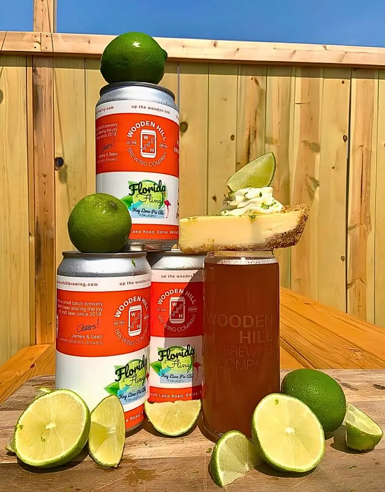Stacked cans of Wooden Hill's Florida Fling Key Lime Pie Ale sit on a sunlit wooden table beside a full glass creatively topped with a slice of pie and surrounded by fresh limes.