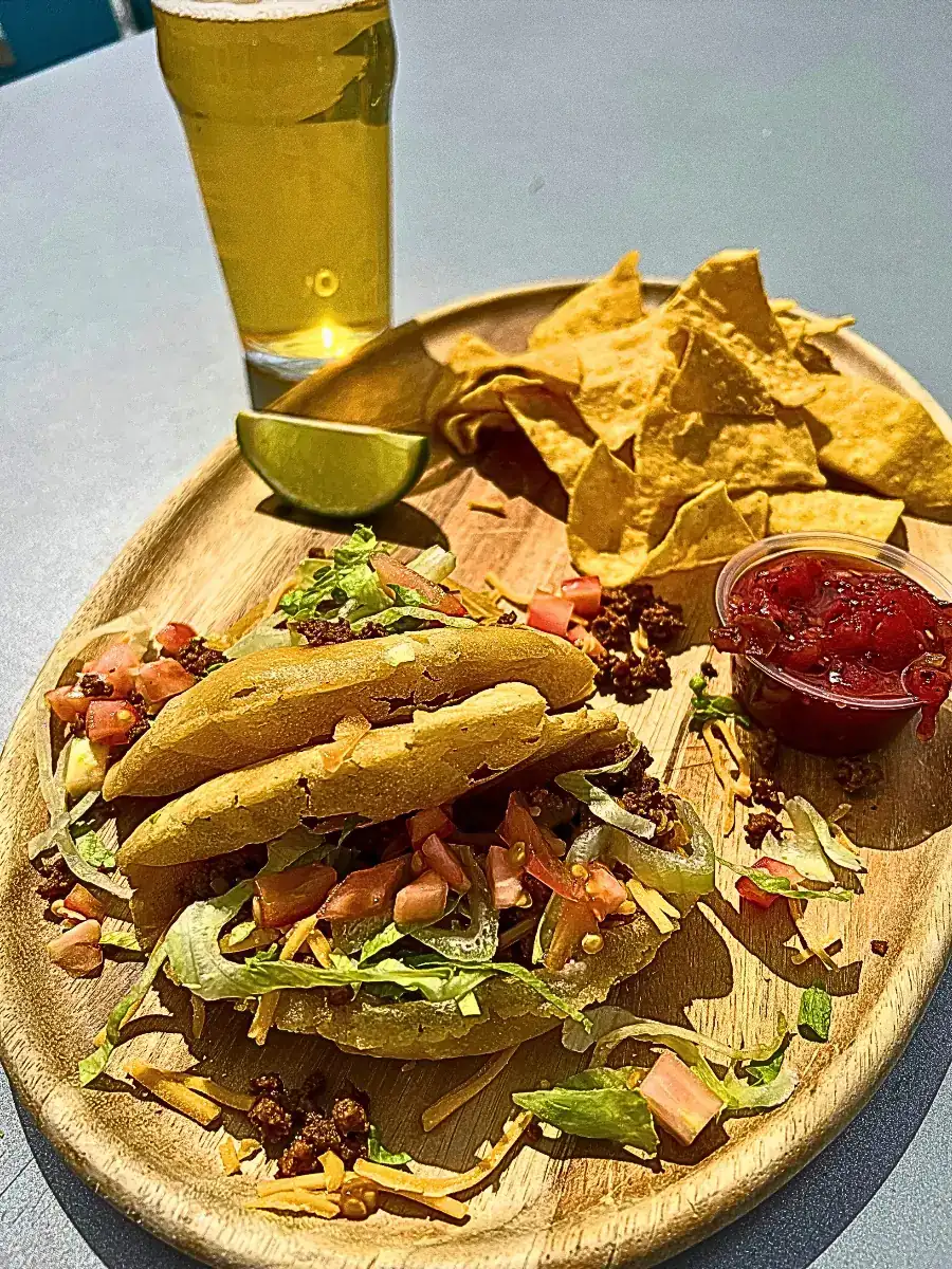 A wooden tray holds two puffy tacos loaded with seasoned ground beef, cheese, lettuce, and tomato, served alongside tortilla chips, salsa, a lime wedge, and a glass of Wooden Hill beer in the background