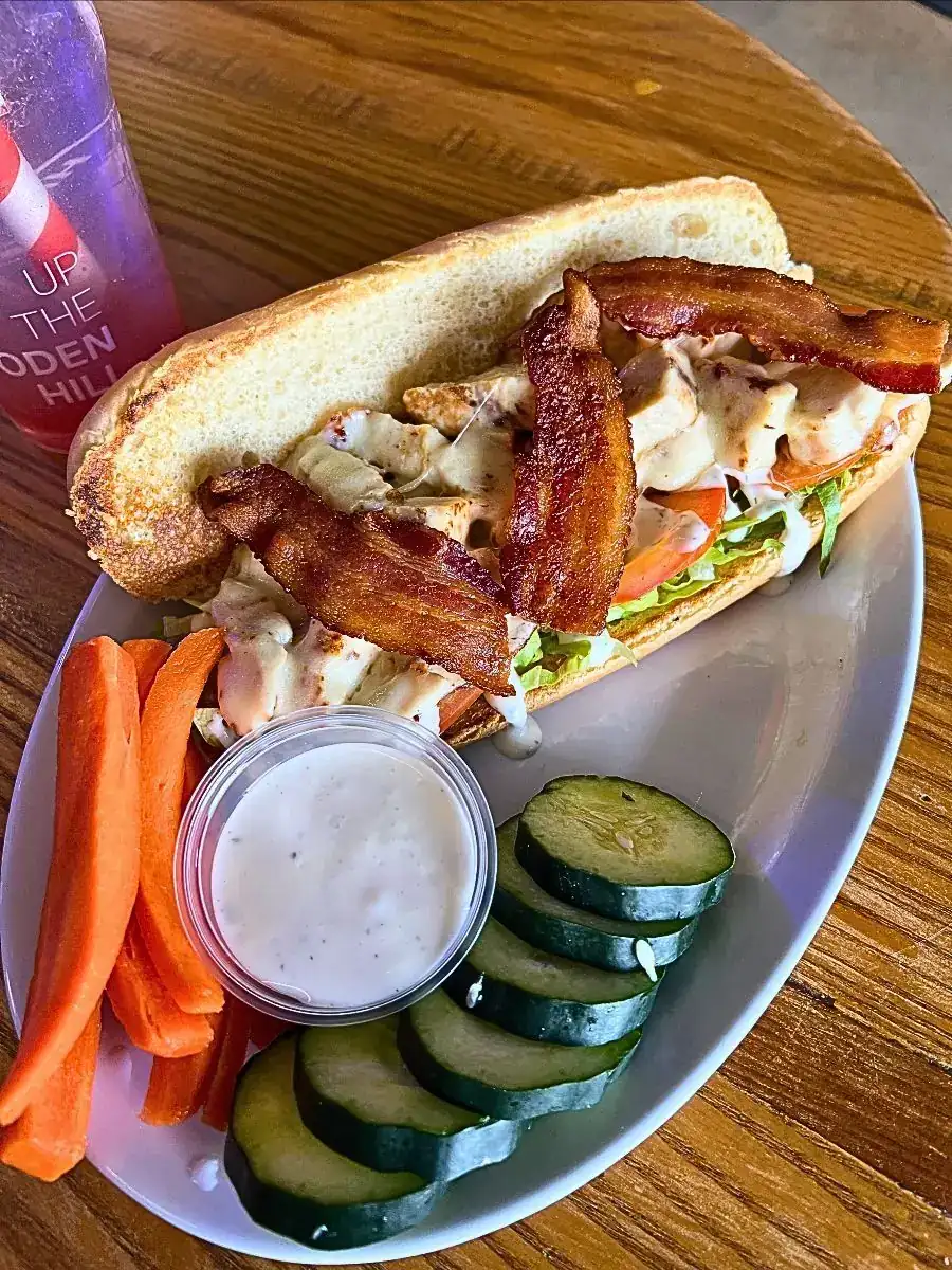 A hearty CBR Philly sandwich loaded with chopped grilled chicken, thick bacon, provolone, fresh veggies, and ranch on a toasted hoagie roll sits on a white plate alongside fresh carrot sticks, cucumber slices, and a side of dip.