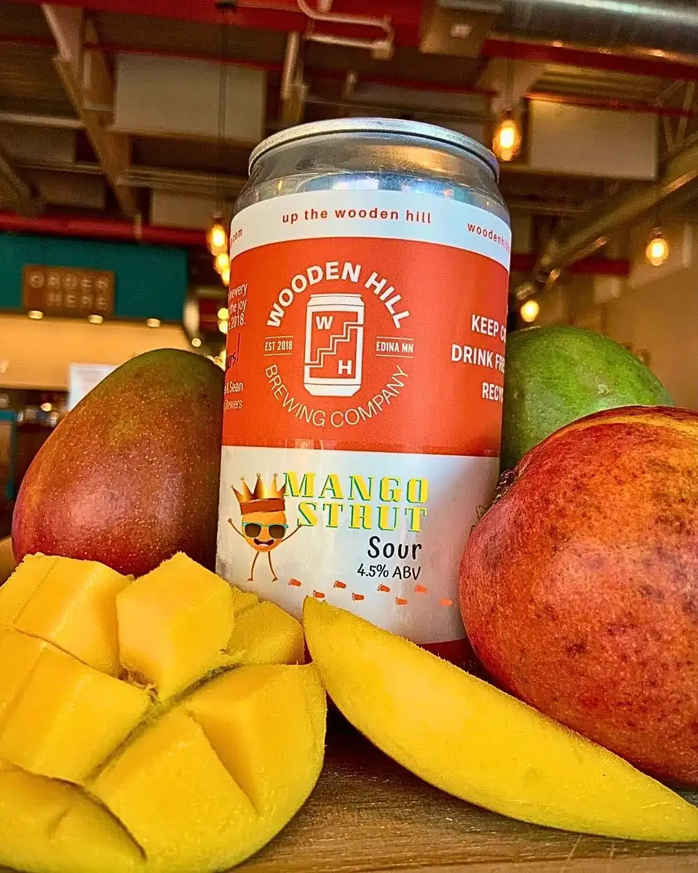 Releasing Wednesday May 20th A can of Wooden Hill Mango Strut Sour Ale surrounded by fresh, sliced, and whole mangoes on a wooden surface, with the warmly lit brewery taproom in the background.