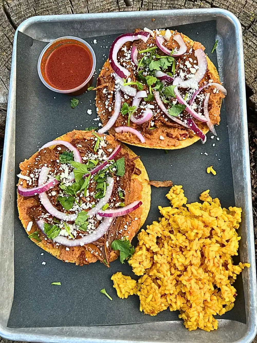 Releasing May 20th, Wednesday Taco Special: A tray holding two crunchy corn tostadas topped with refried beans, shredded birria beef, red onion, queso fresco, and cilantro. Served with a side of Spanish rice and a small cup of consommé dipping sauce.