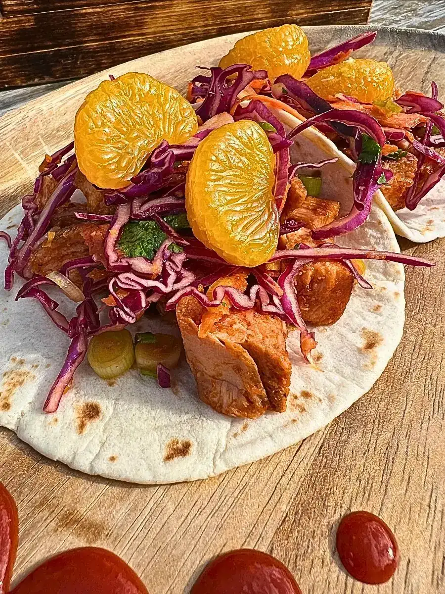 Available starting Wednesday, May 27th.Garlic Sriracha Chicken Tacos- Two soft flour tacos filled with grilled chicken, red cabbage slaw, green onions, and mandarin oranges on a wooden board.
