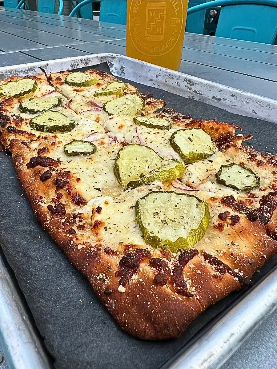 Available starting Thursday, May 28th. Pickle Maxxer Flatbread- A rectangular flatbread topped with melted mozzarella, pickle chips, sliced red onions, and herbs on a metal tray, with a glass of beer in the background.