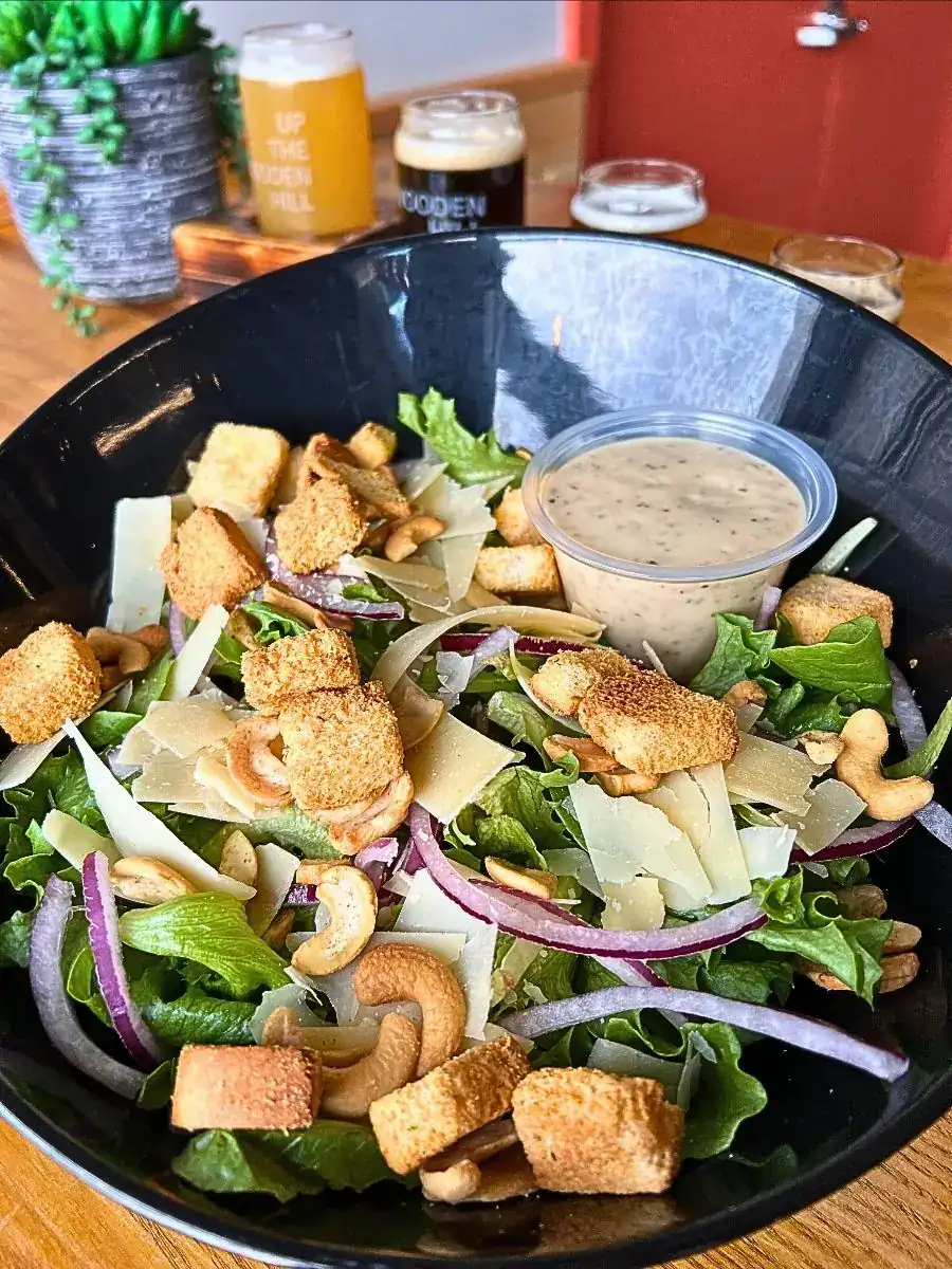 A black bowl filled with a fresh Caesar Salad featuring mixed greens, shaved Parmesan cheese, house rosemary cashews, red onions, and croutons. Served with a side of creamy dressing on a wooden table with a beer flight in the background.