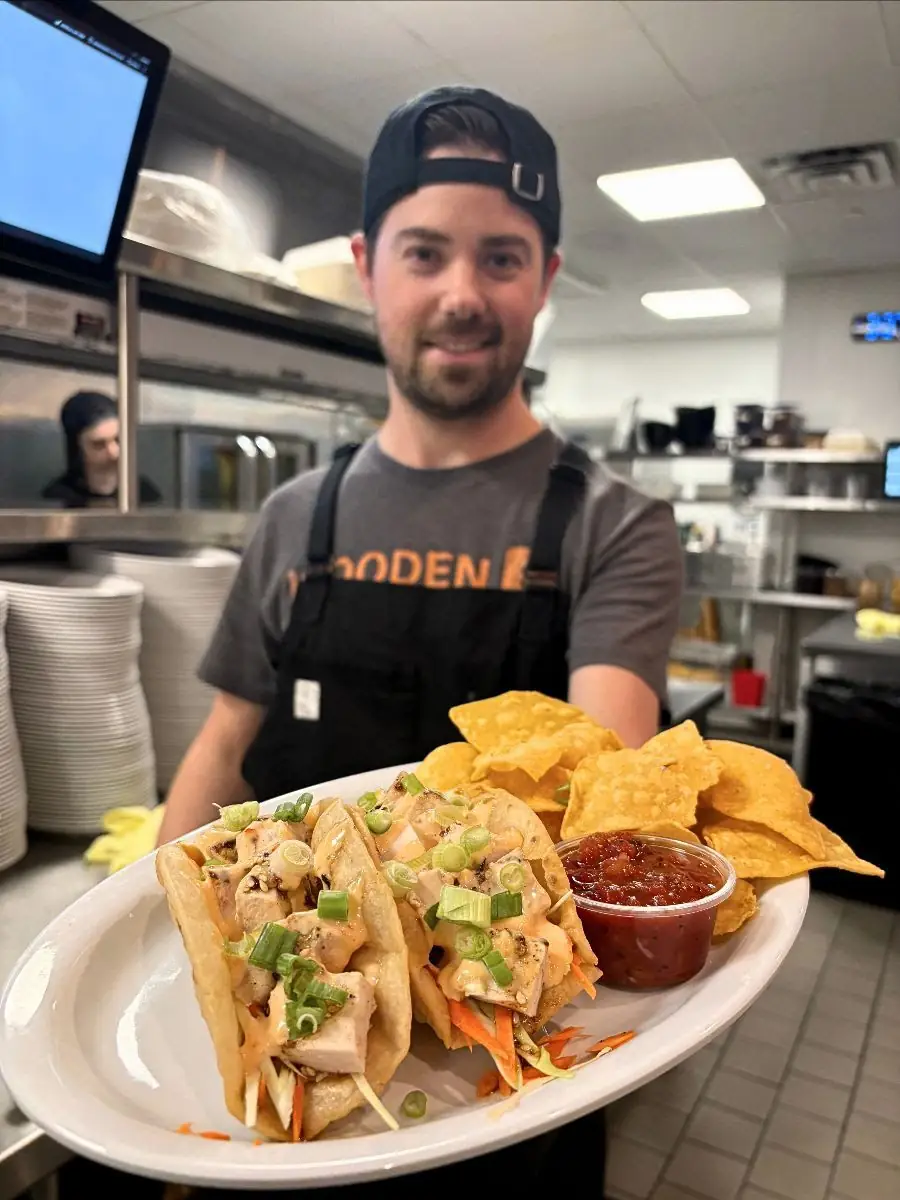 Image of a guy in the kitchen holding a plate out in front of him with two egg roll tacos, chips, and salsa on the plate.