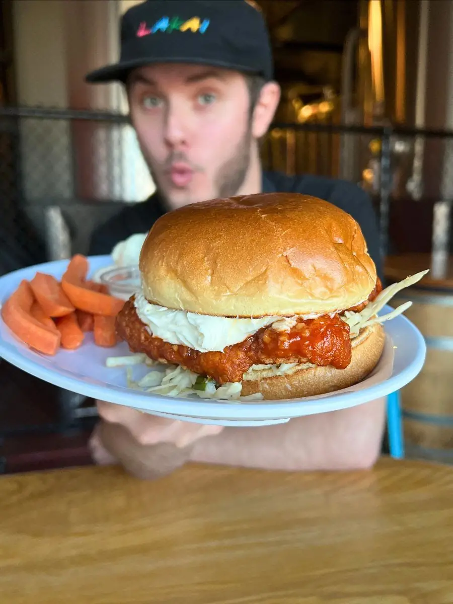 Image of a guy holding a plate out in front of him. Focus is on a Garlic Sriracha Chicken sandwich with carrots on the plate in the backgroud.