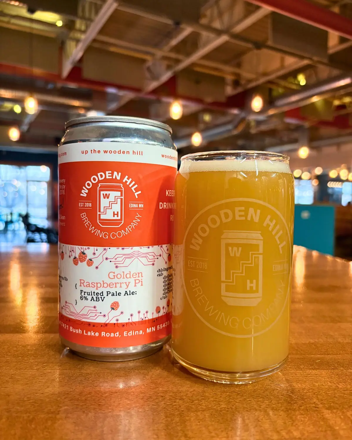 Image of Wooden Hill beer can of Golden Raspberry Pi on the left and a Wooden Hill glass with a hazy beer on the right with the inside of the brewery in the background.