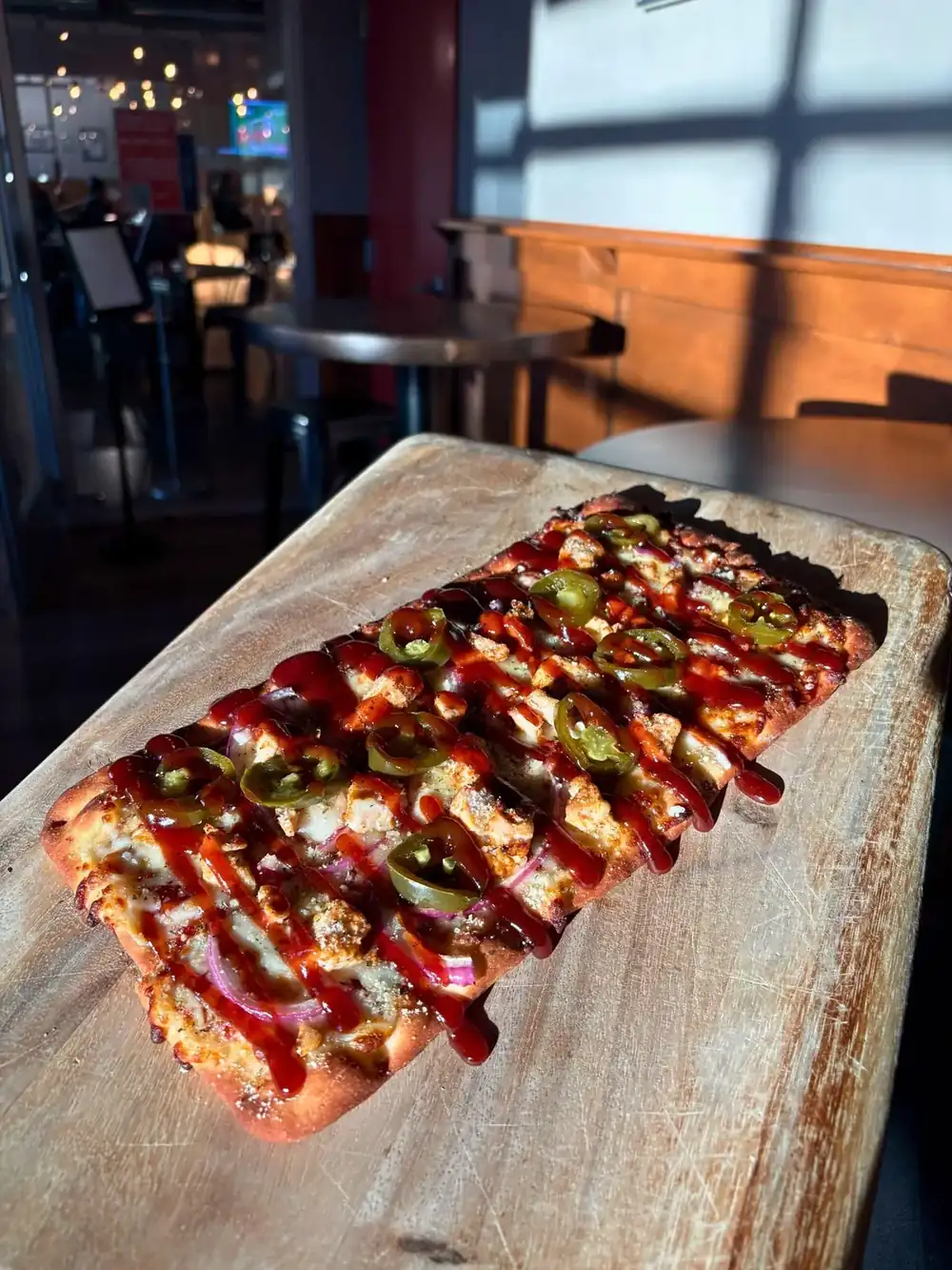 Image of BBQ Chicken flatbread on a wood board with the inside of the brewery in the background.