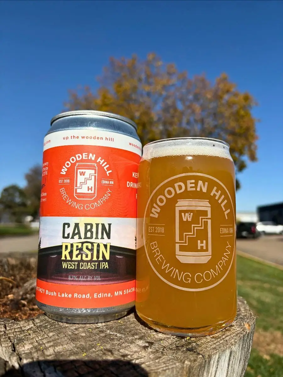 Image of Wooden Hill can on the left with Cabin Resin logo. Wooden Hill branded glass with an IPA on the right. On a tree stump with trees and leaves in the background.