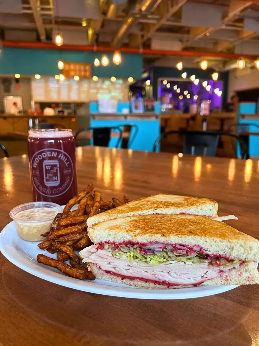 Image of the gobbler sandwich with sweet potato fries and a dipping container on the plate. Wooden Hill branded glass with a berry colored beer in it. Brewery in the background.