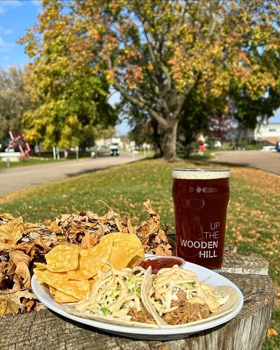 Plate with two pigs in a pumpkin tacos on it with chips and salsa. Wooden Hill branded glass with a darker beer in the background. All on a tree stump with leaves and grass in the background.