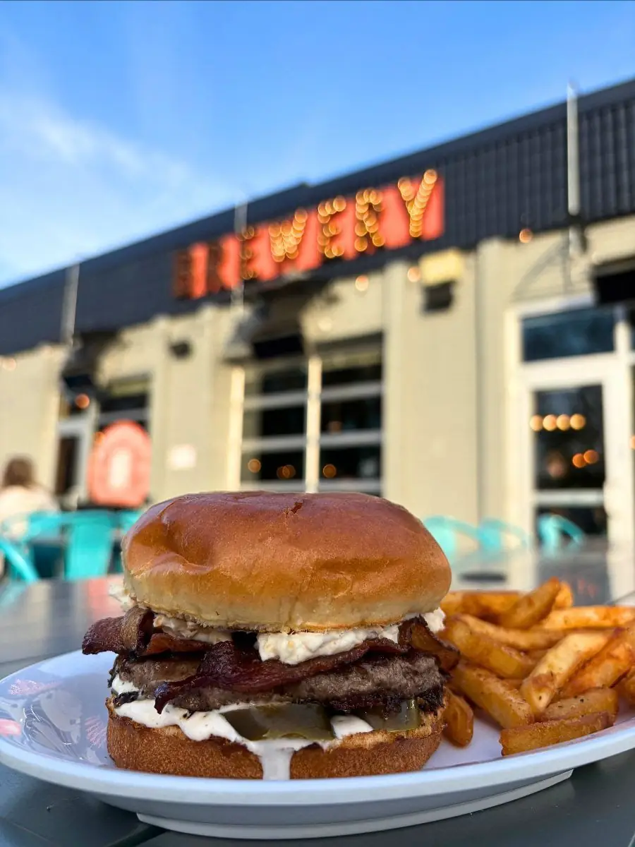 Image focused on a jalapeno popper burger with fries on a plate. Background is of the front of Wooden Hill with the Brewery sign lit up.