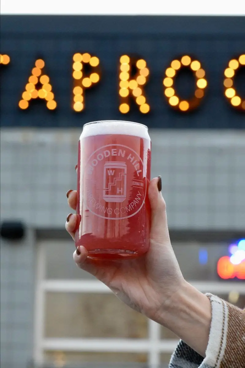 Photo of pixie catcher sour beer in a Wooden Hill branded glass, held up in front of outside light up taproom sign