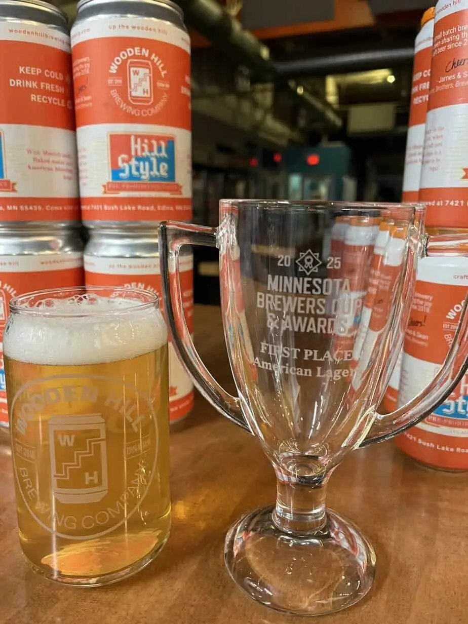 photograph of a glass cup trophy for best american lager, next to a pint and cans of the winning beer, "hill style."