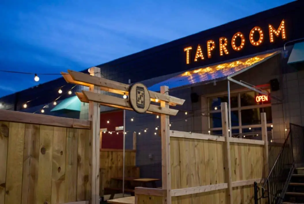Photo of the Wooden Hill Brewing taproom at twilight, featuring the lit "taproom" sign and the entrance to the fenced patio.