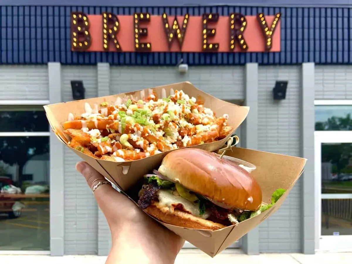 A photo of a hand holding two baskets of food, one full of french fries and one with a gourmet burger - in front of the "brewery' sign outside of the Wooden Hill taproom.